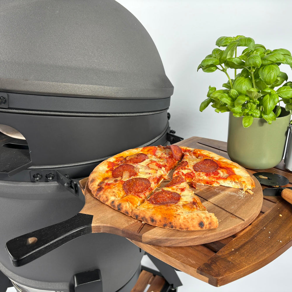Pizza on a wooden serving board beside an Off-Black kamado and a basil plant.