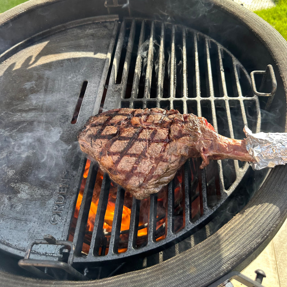 Bone-in steak searing on a half-moon grate over flames inside a kamado.