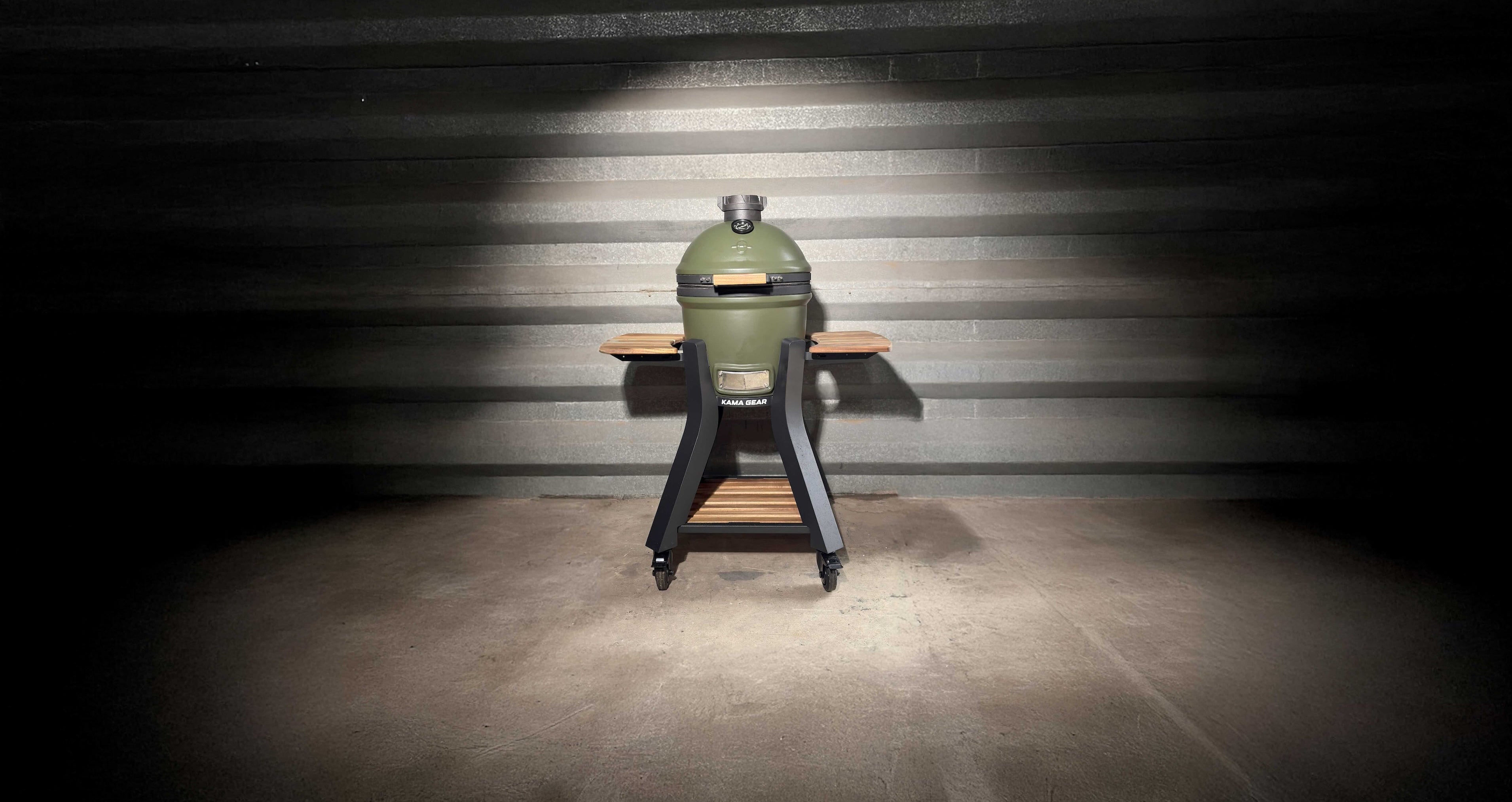 olive green Kamado barbecue grill on a stand against a dark, striped wall.